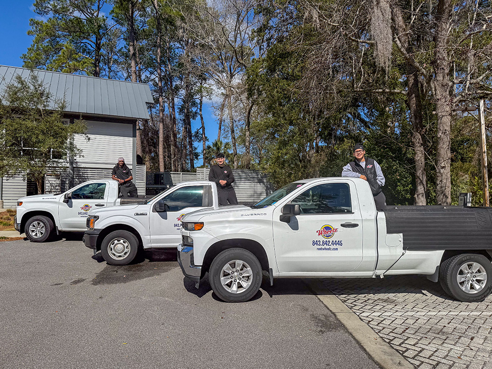 Three people standing on white trucks with logos outside a building, surrounded by trees.